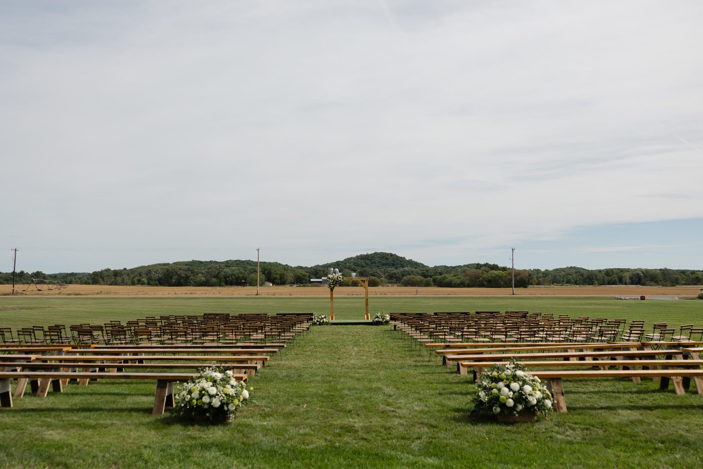 An open-field ceremony set up before guests arrive, with wooden benches and chairs lining a grass aisle to a flower-topped wooden arch