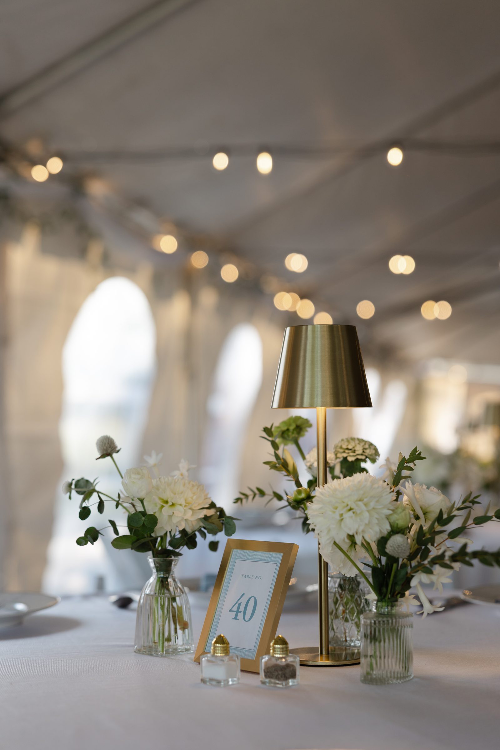 A reception table under a tent with white flowers in glass bud vases, a brass lamp, and a framed table number beneath string lights