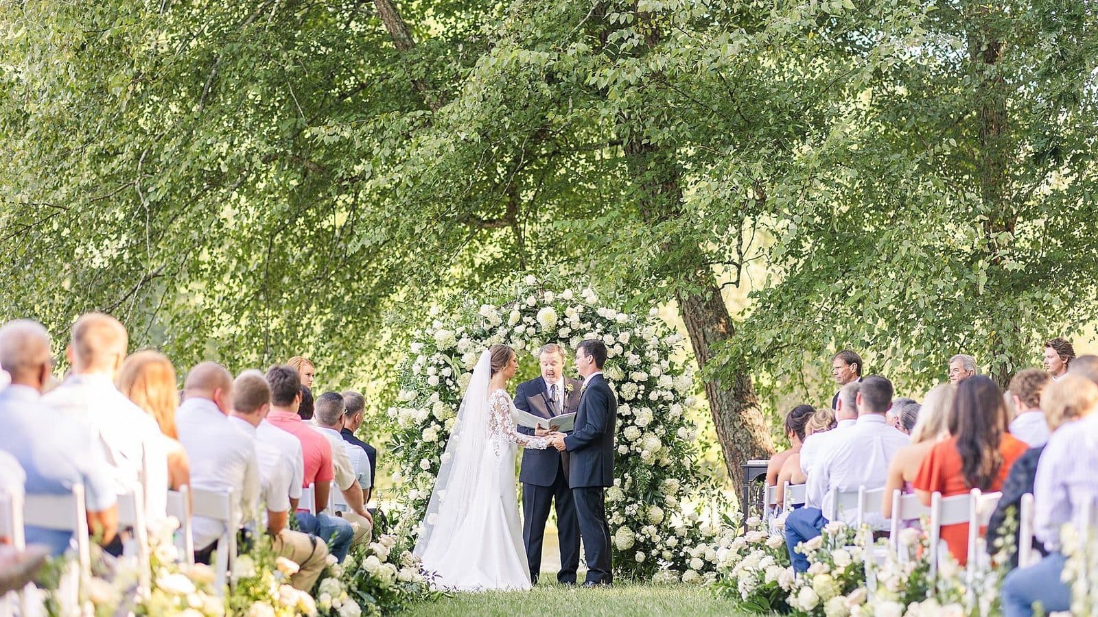 Outdoor wedding ceremony under large trees at Heyward Manor, photographed by Katie Jaynes Photography